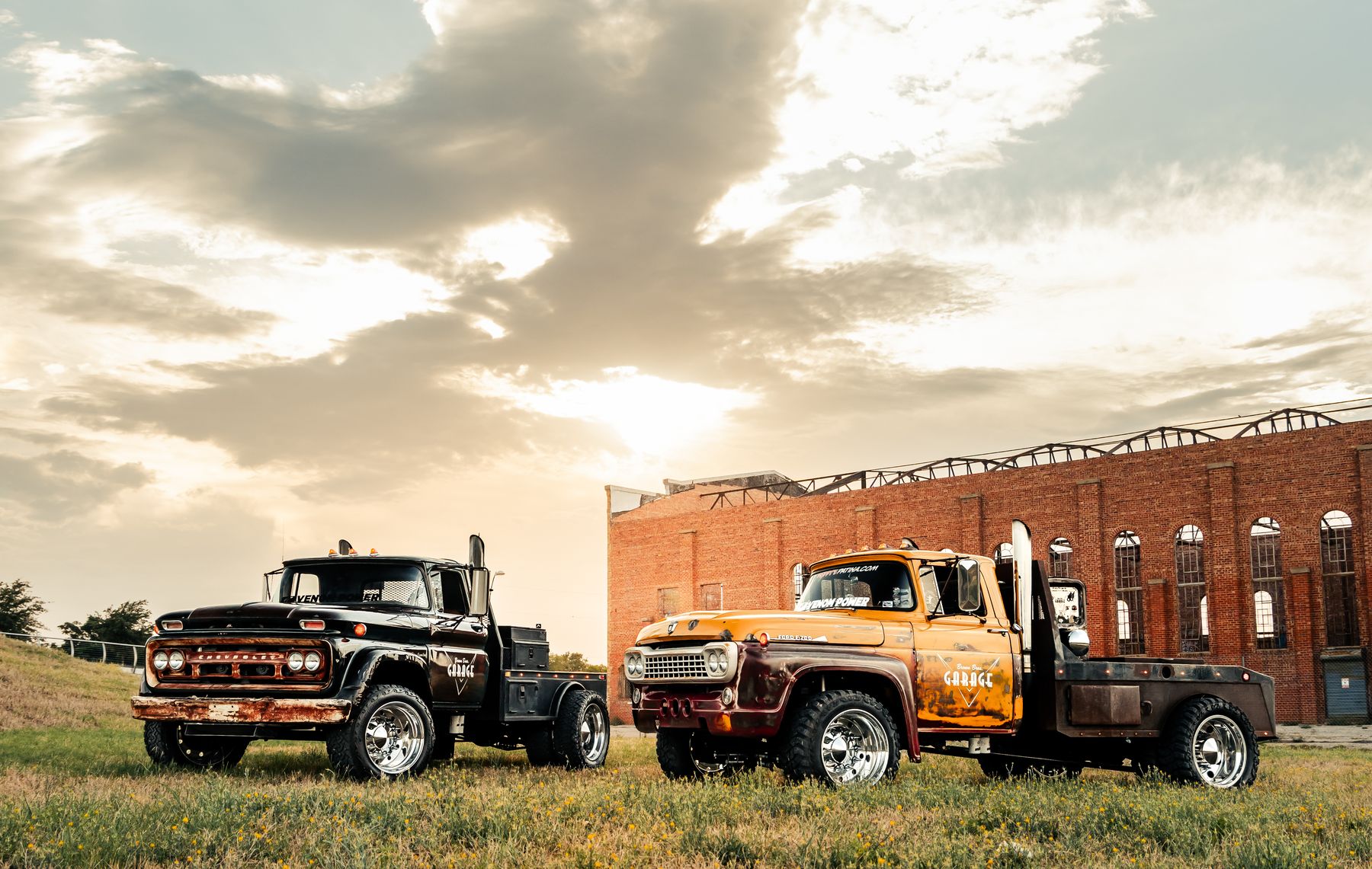 Two restored diesel work trucks, one black and one orange, parked on grass in front of a historic brick building at sunset
