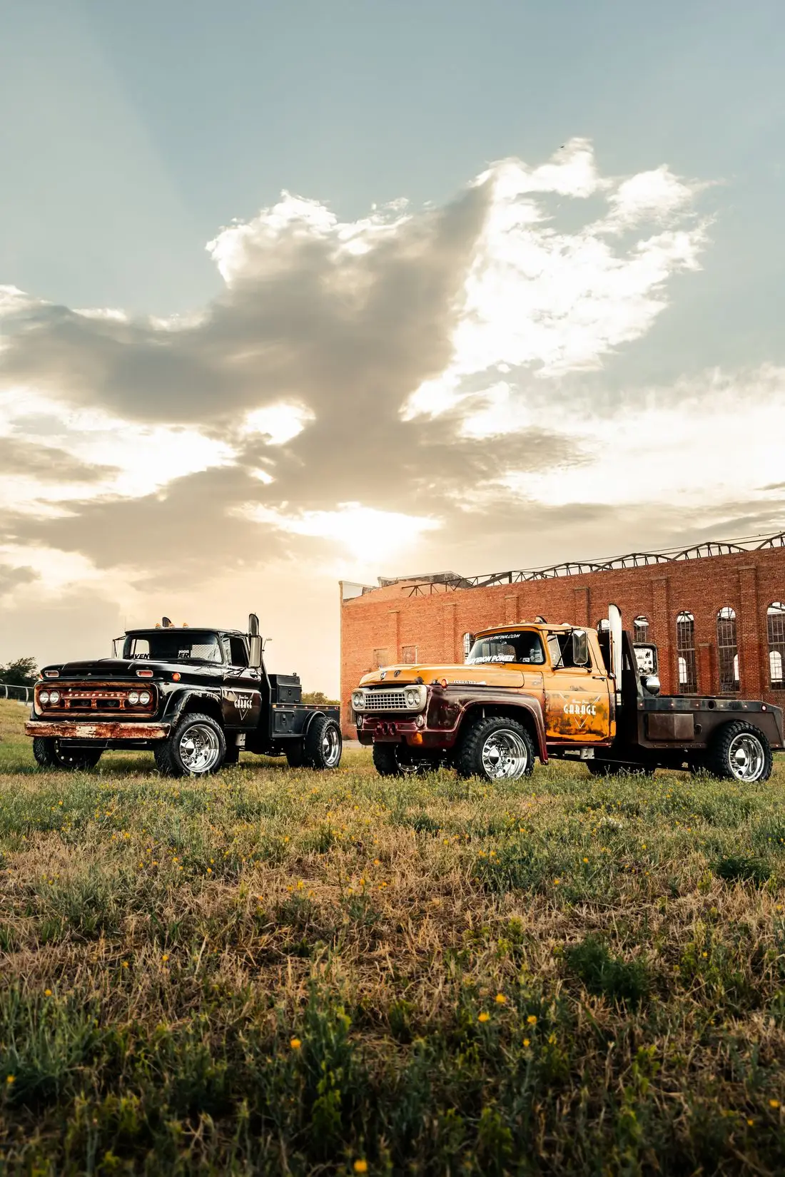 Two restored diesel work trucks, one black and one orange, parked on grass in front of a historic brick building at sunset