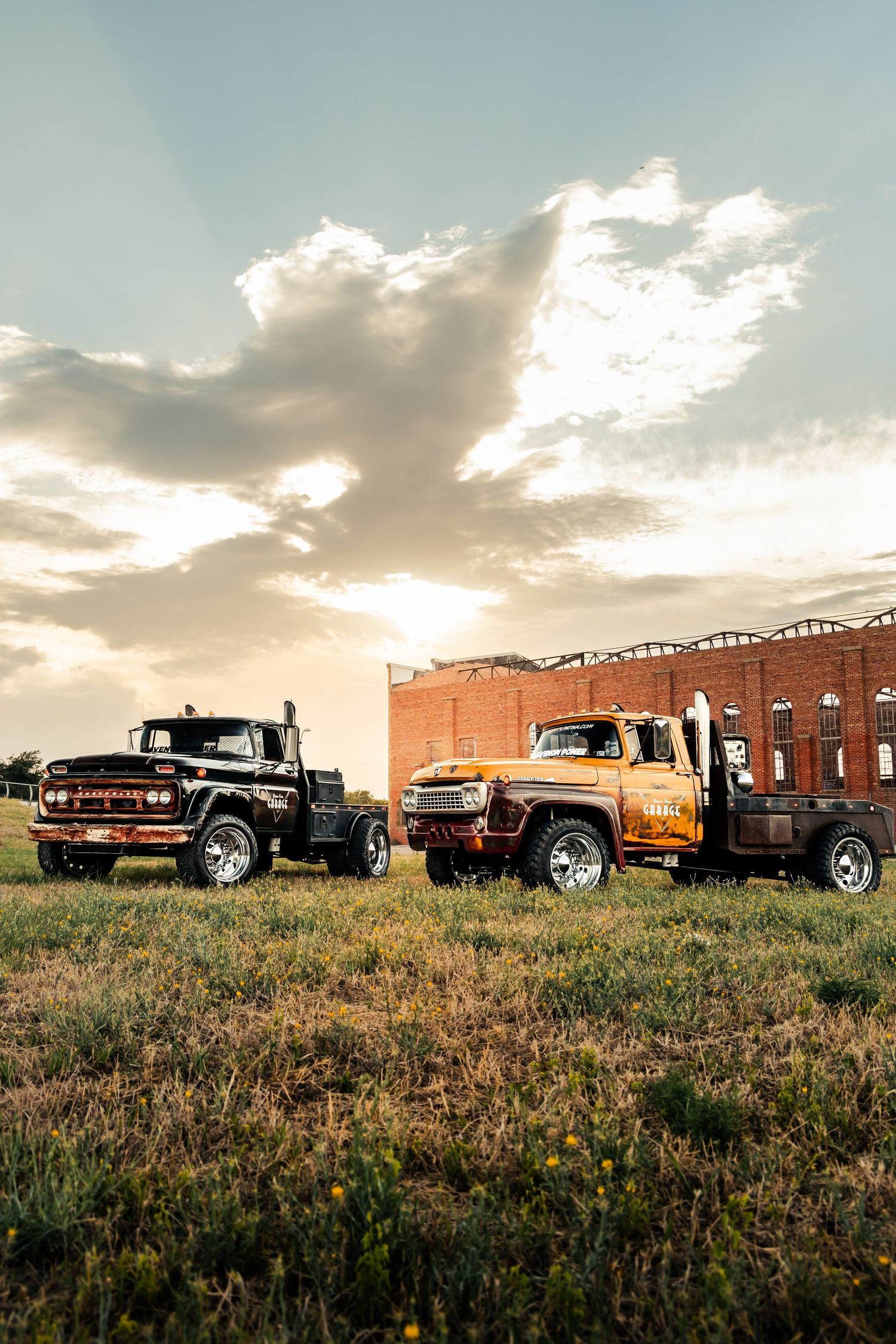 Two restored diesel work trucks, one black and one orange, parked on grass in front of a historic brick building at sunset