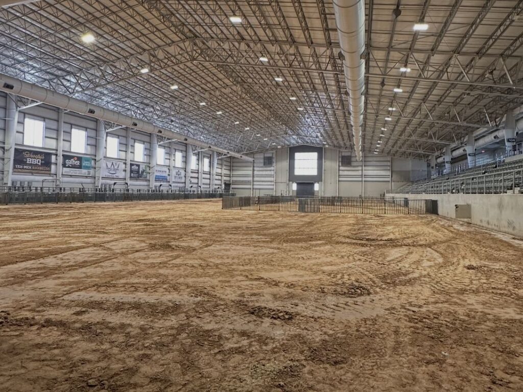 The empty Equine Complex Arena with a dirt floor, stadium seating and a large steel roof